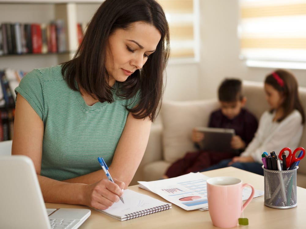 Mãe trabalhando em sua mesa de trabalho organizada, enquanto os filhos assistem no Ipad desenhos animados bem comportados.