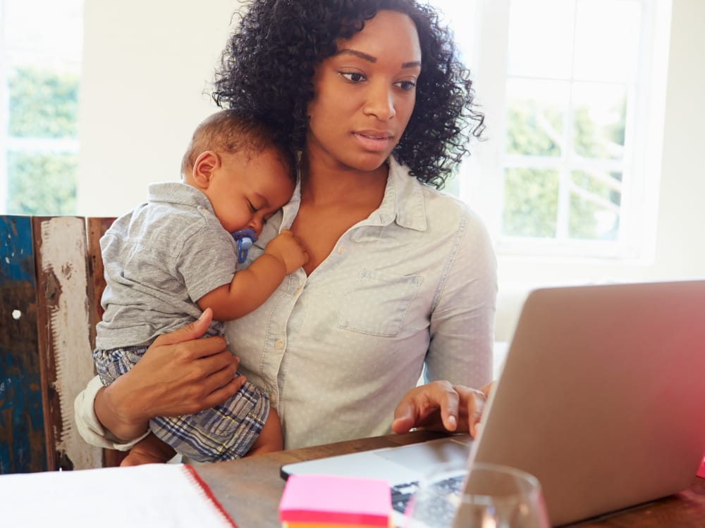 Mãe trabalhando em casa no computador enquanto o bebê dorme em seu colo.