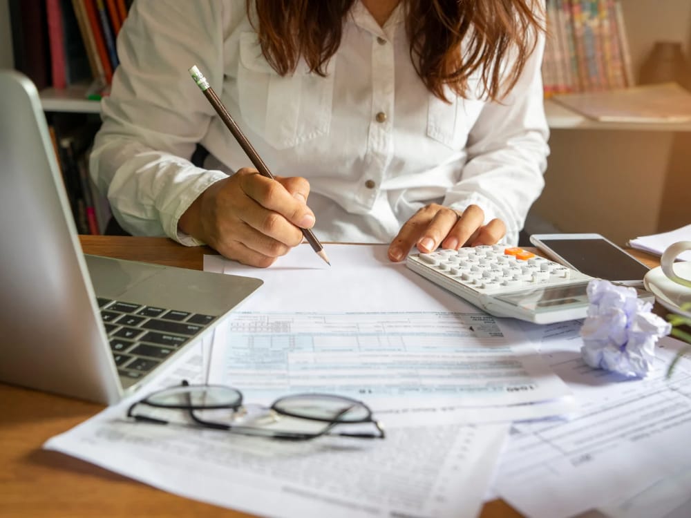 Uma mulher está sentada em uma mesa com um caderno, uma calculadora e planilhas de orçamento. Ela é focada e determinada ao definir metas financeiras e planos para o futuro